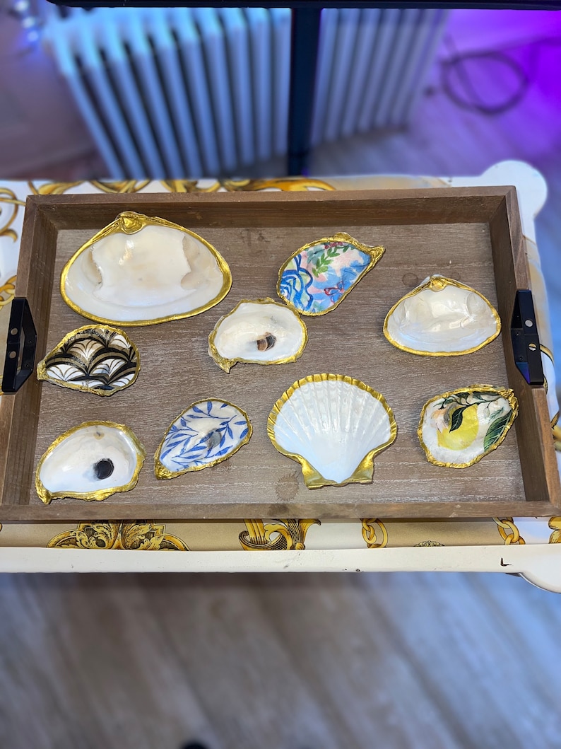 May include: A wooden tray with nine decorative seashells. The shells are painted with various patterns and designs, including floral, geometric, and abstract motifs. The shells are arranged in a circular pattern on the tray, with a gold rim around each shell.