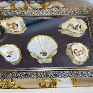 May include: A silver tray with a mirrored surface and ornate gold trim. Five white seashell-shaped dishes with gold accents hold various jewelry pieces, including pearl earrings and a gold necklace with a pendant.