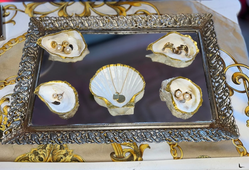 May include: A silver tray with a mirrored surface and ornate gold trim. Five white seashell-shaped dishes with gold accents hold various jewelry pieces, including pearl earrings and a gold necklace with a pendant.