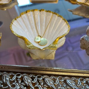 May include: A gold necklace with a small square pendant and a larger round pendant, resting in a white seashell dish with a gold rim. The dish is on a silver tray with a decorative floral design.