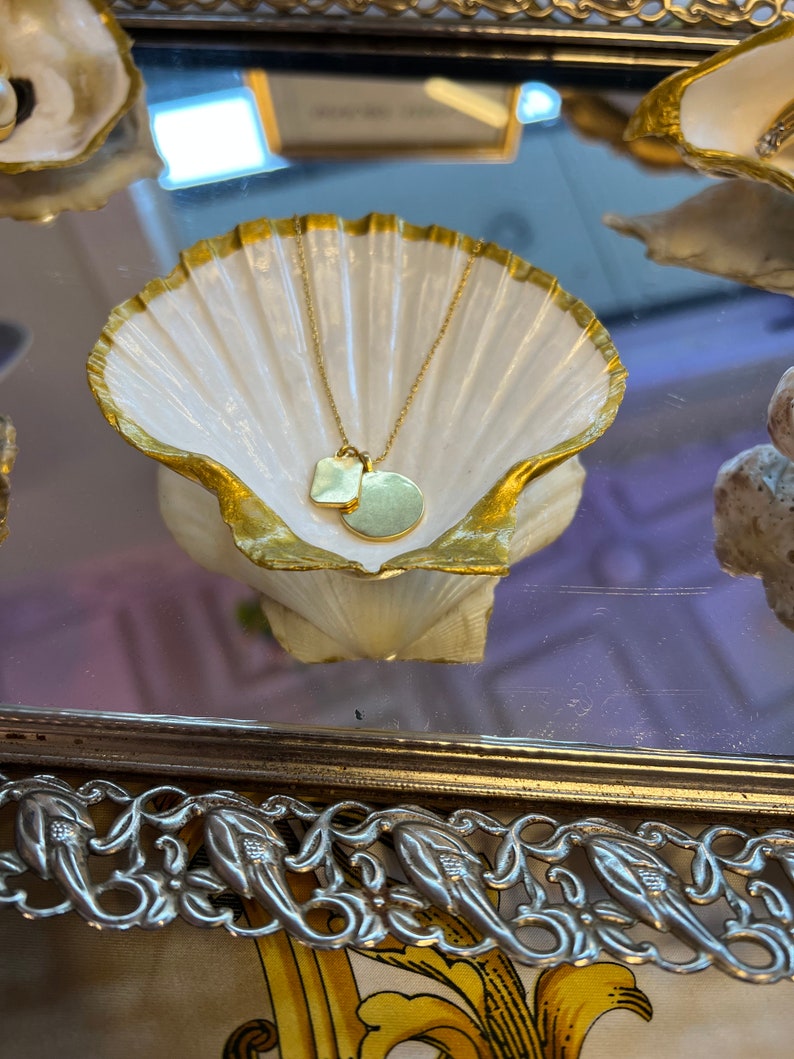 May include: A gold necklace with a small square pendant and a larger round pendant, resting in a white seashell dish with a gold rim. The dish is on a silver tray with a decorative floral design.