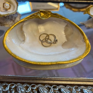 May include: A white seashell ring dish with a gold rim. The dish is filled with three gold rings.