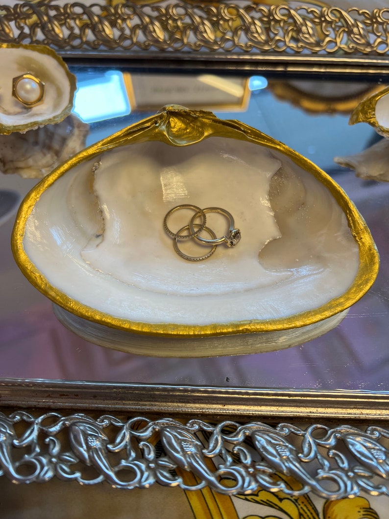 May include: A white seashell ring dish with a gold rim. The dish is filled with three gold rings.