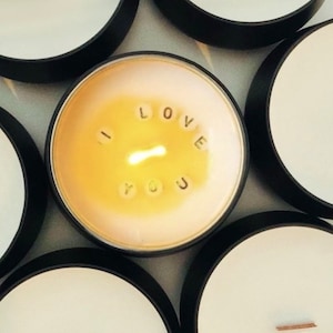 May include: A round candle in a black metal tin with white wax. The wax is partially melted, revealing small white letter beads arranged in a circle around the central wick that spell out the phrase I LOVE YOU in black capital letters.