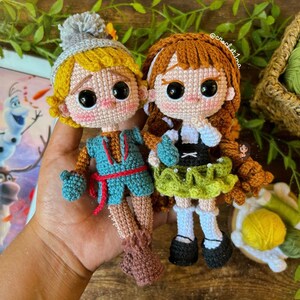 May include: Two crocheted dolls, one with brown hair and a green dress, the other with blonde hair and a blue and red outfit. The dolls are holding hands and have large, black eyes.