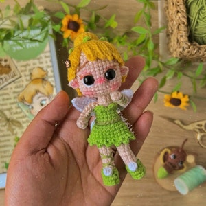 Puede incluir: Una muñeca de crochet de Campanilla con vestido verde, cabello amarillo y grandes ojos negros. La muñeca sostiene una pequeña flor verde en su mano.