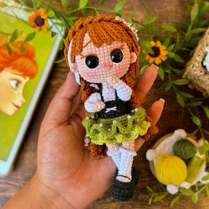 May include: Crocheted doll with brown hair, wearing a white and green dress with a black belt. The doll is holding her hands to her face and has large black eyes. The doll is sitting in a hand.