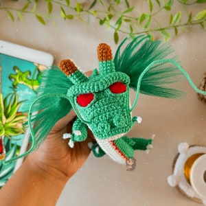 Puede incluir: Dragón de crochet verde con ojos rojos y cuerpo blanco y rojo. El dragón tiene un largo pelo de lana verde.