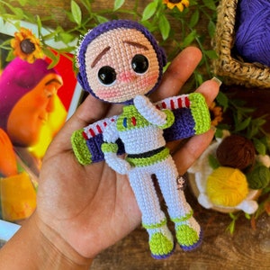 Puede incluir: Un juguete de crochet de Buzz Lightyear de la franquicia Toy Story. El juguete lleva un traje espacial blanco con detalles verdes y rojos. El juguete tiene una cabeza morada con una expresión triste y grandes ojos negros.