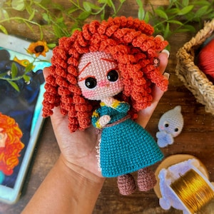 Puede incluir: Una muñeca de crochet con pelo rojo, un vestido azul y botas marrones. La muñeca sostiene una pequeña flor azul y blanca de crochet.