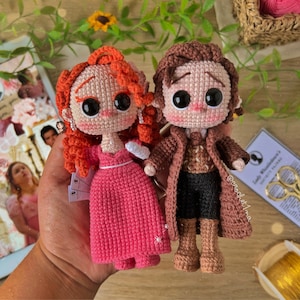 Pode incluir: Duas bonecas de crochê, uma com cabelo ruivo e um vestido rosa, a outra com cabelo castanho e um casaco marrom. As bonecas estão em pé em uma mão.