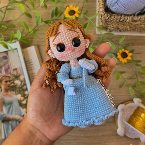 Pode incluir: Uma boneca de crochê com cabelo castanho, olhos grandes e um vestido azul. A boneca está usando um cinto dourado e um colar azul. A boneca está sendo segurada em uma mão.