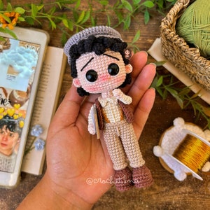 May include: A handmade crochet doll of a young man, wearing a gray cap, brown vest, and tan pants. The doll has large black eyes and is holding a small book. The doll is approximately 5 inches tall.