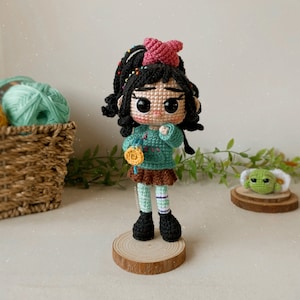 May include: A crocheted doll with black hair, a pink bow, and a green sweater. The doll is holding a yellow flower and is standing on a wooden base.