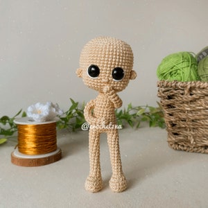 Puede incluir: Una muñeca beige tejida a ganchillo con grandes ojos negros y una pose pensativa. La muñeca tiene una cabeza redonda, orejas pequeñas y un cuerpo simple. El nombre del creador de la muñeca, "@crochetina", es visible.