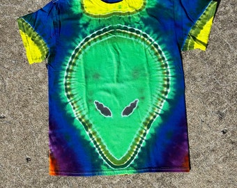 May include: A tie-dye t-shirt with a green alien face design. The shirt is blue, green, yellow, and purple.