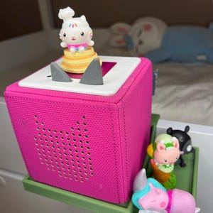 Puede incluir: Un altavoz en forma de cubo rosa brillante con una parte superior blanca, detalles grises y una bandeja verde. Una figura de gato blanco con un gorro de chef se encuentra encima del altavoz. Figuras coloridas adicionales, incluyendo un cerdo y un oso negro, están en la bandeja.