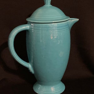 May include: A turquoise ceramic coffee pot with a lid and handle. The pot has a rounded body, a pedestal base, and a curved handle. The lid has a small knob on top. The pot is a solid turquoise color.