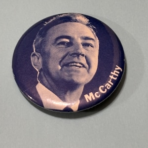 May include: A round, navy blue button featuring a portrait of a smiling man in a suit. The name "McCarthy" is printed in white along the bottom edge of the button. The image is set against a light blue background.