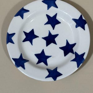 May include: A round white ceramic plate featuring a scattered pattern of hand-painted dark blue stars. The stars vary slightly in size and orientation, covering both the center and the rim of the plate, creating a simple and uniform design.