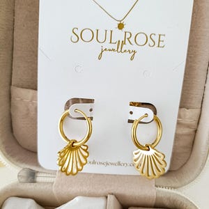 Puede incluir: Pendientes de aro dorados con un dije de concha. Los pendientes se exhiben en una tarjeta blanca con el texto "SOUL ROSE jewellery" en letras doradas. También se ve un collar dorado.