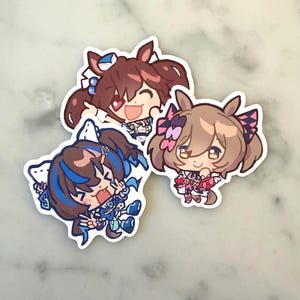 Op de afbeelding: Drie stickers in anime-stijl met personages met grote ogen en kleurrijk haar. De stickers hebben een witte rand en staan in verschillende poses, met één glimlachend en twee met opgewonden uitdrukkingen. De personages dragen gedetailleerde outfits.