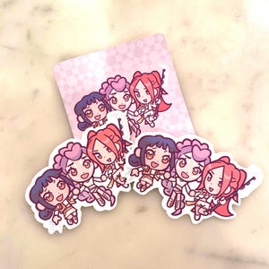 Op de afbeelding: Een verzameling stickers met anime-meisjes met verschillende kapsels en outfits. De stickers hebben een witte rand en zijn gerangschikt op een lichtroze achtergrond. De meisjes hebben verschillende haarkleuren, waaronder rood, paars en donkerblauw.