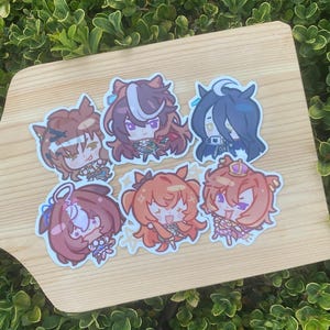 Può includere: Una collezione di sette adesivi in stile anime con personaggi colorati con capelli e abiti dettagliati. Gli adesivi sono disposti su una superficie di legno chiaro, su uno sfondo di fogliame verde.