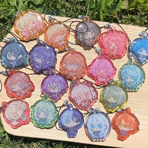 May include: A collection of colorful anime-style character charms. Each charm features a unique character design with varying hair colors, outfits, and expressions. The charms are attached to black cords and are displayed on a wooden surface.