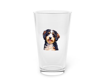 Puede incluir: Un vaso de pinta de vidrio transparente con una ilustración colorida de un perro Bernedoodle en la parte delantera.