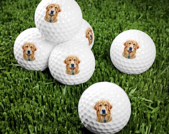 May include: Six white golf balls with a printed image of a golden retriever dog on each ball. The golf balls are arranged in a semi-circle on a green grassy surface.