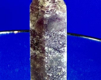 May include: A purple crystal point with a faceted shape. The crystal has a textured surface with white and grey inclusions. The crystal is standing upright against a blue background.