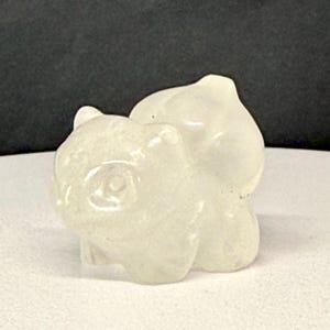 May include: A white, opaque, ceramic figurine of a small animal, possibly a pig or a bear, curled up in a sleeping position.