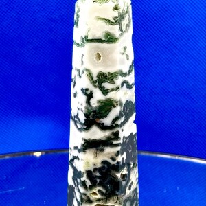 May include: A tall, polished moss agate obelisk. The stone features a mix of white, black, and green patterns. The top is a pointed pyramid shape. The obelisk is a decorative item.