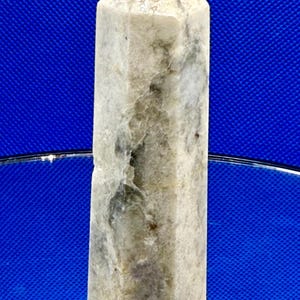 May include: A tall, rectangular crystal obelisk with a pointed top. The stone has a mottled appearance with shades of gray, white, and hints of green. The obelisk is standing upright, reflecting on a mirrored surface against a blue background.