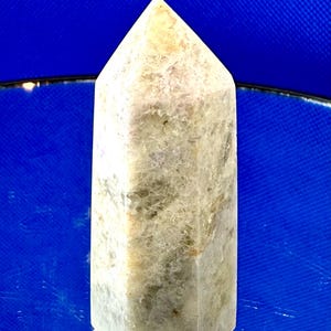 May include: A light-colored, rectangular crystal obelisk with a pointed top. The crystal has a textured surface with shades of beige, gray, and green. The obelisk is displayed on a reflective surface against a blue background.