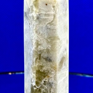 May include: A tall, rectangular crystal obelisk with a pointed top. The stone is primarily off-white with areas of light green and brown. The obelisk is set against a vibrant blue background.