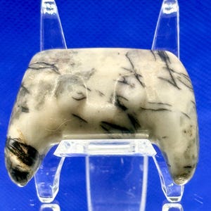 May include: A polished, cream-colored stone pendant with black veining, displayed on a clear acrylic stand. The stone has a rounded top and two curved protrusions at the bottom. The background is a solid blue.