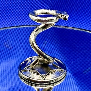 May include: A metallic silver-toned display stand featuring a coiled snake figure rising from a circular base. The base is decorated with a star-like geometric pattern. The entire structure has a textured, reflective finish and a vertical spiral design.