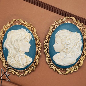 May include: Two gold-toned cameo brooches with blue enamel and white porcelain figures. The figures are in profile and have detailed hair and clothing.