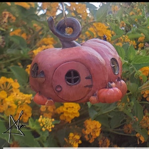 Op de afbeelding: Een kleine, handgeschilderde keramische pompoen met een deur en ramen. De pompoen is geschilderd in tinten rood en bruin en heeft een sprookjesachtige, speelse uitstraling. Hij hangt aan een touwtje en is omringd door gele bloemen.