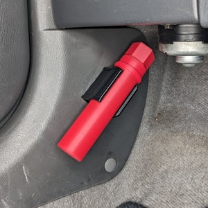 Könnte beinhalten: Ein rotes, zylindrisches Sicherheitsgerät, möglicherweise eine Straßenfackel, befestigt durch einen schwarzen Clip. Das Gerät befindet sich in der Nähe des Fahrzeugbodens, an einer grauen Kunststoffplatte und einem grauen Teppich.