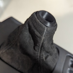 Pode incluir: Botão de cambio de camurça preta com parte superior de plástico preto. O botão está instalado no console central de um carro.