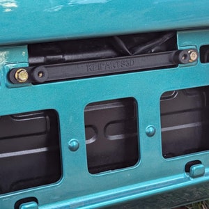 May include: A teal car bumper with a black licence plate holder. The holder has three rectangular cutouts and the text "KEIFANTS 3D" printed on it. There are four silver screws holding the holder in place.