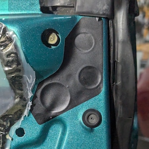 May include: Close-up of a teal car door frame with black plastic components. Visible are a bolt, a rubber grommet, and various circular indentations. The door frame has a metallic finish.