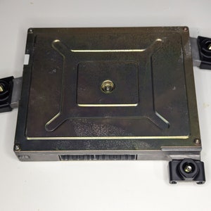 Peut inclure: Unité de commande électronique de couleur argentée avec des supports de montage en plastique noir. L'unité a une forme rectangulaire et une surface texturée.