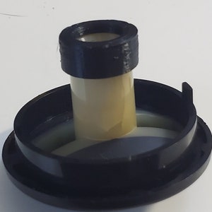 May include: A black and beige plastic object with a cylindrical center and a wide, circular base. The center has a smaller, raised black ring at the top. The object appears to be a component or part of a larger mechanism.