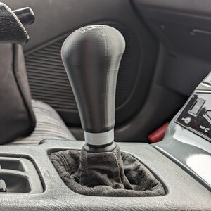 May include: A black leather-wrapped gear shift knob with a white stripe and a black plastic base. The knob is installed in a car's center console.