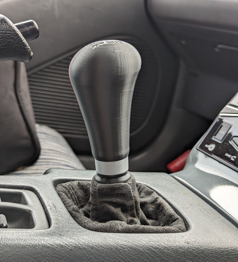 May include: A black leather-wrapped gear shift knob with a white stripe and a black plastic base. The knob is installed in a car's center console.