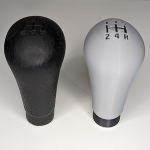 May include: Two gear shift knobs, one black and one white. The black knob has a textured surface and the white knob is smooth. Both knobs have a gear shift pattern on the top.